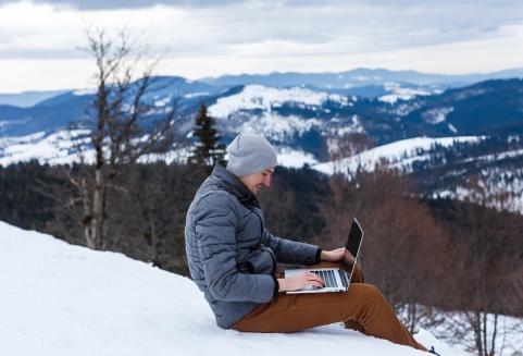 Nuori aikuinen istuu vuorimaisemassa lumisessa mäessä tietokoneen ääressä.