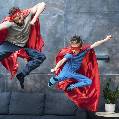 Isä ja poika hyppäävät sohvalta supermiesasut yllä.