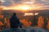 Ihminen katselee auringonnousua korkealla kalliolla. Ruskapuita ja järvi.