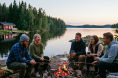 Viisi ihmistä istuu nuotiolla paistamassa makkaraa valoisassa kesäyössä. Taustalla järvi ja punainen kesämökki.